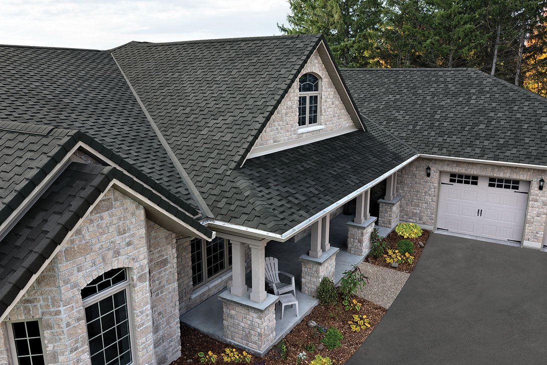A large house with light stone walls and dark gray asphalt shingle roofing by Exterior Done Best, featuring multiple gables, a covered porch with chairs, and a paved driveway next to landscaped flower beds in the Twin Cities.