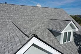 A close-up view of a house roof covered with gray asphalt shingles, showcasing expert roofing by Exterior Done Best in the Twin Cities, with multiple roof angles and a dormer with large windows under a clear blue sky.
