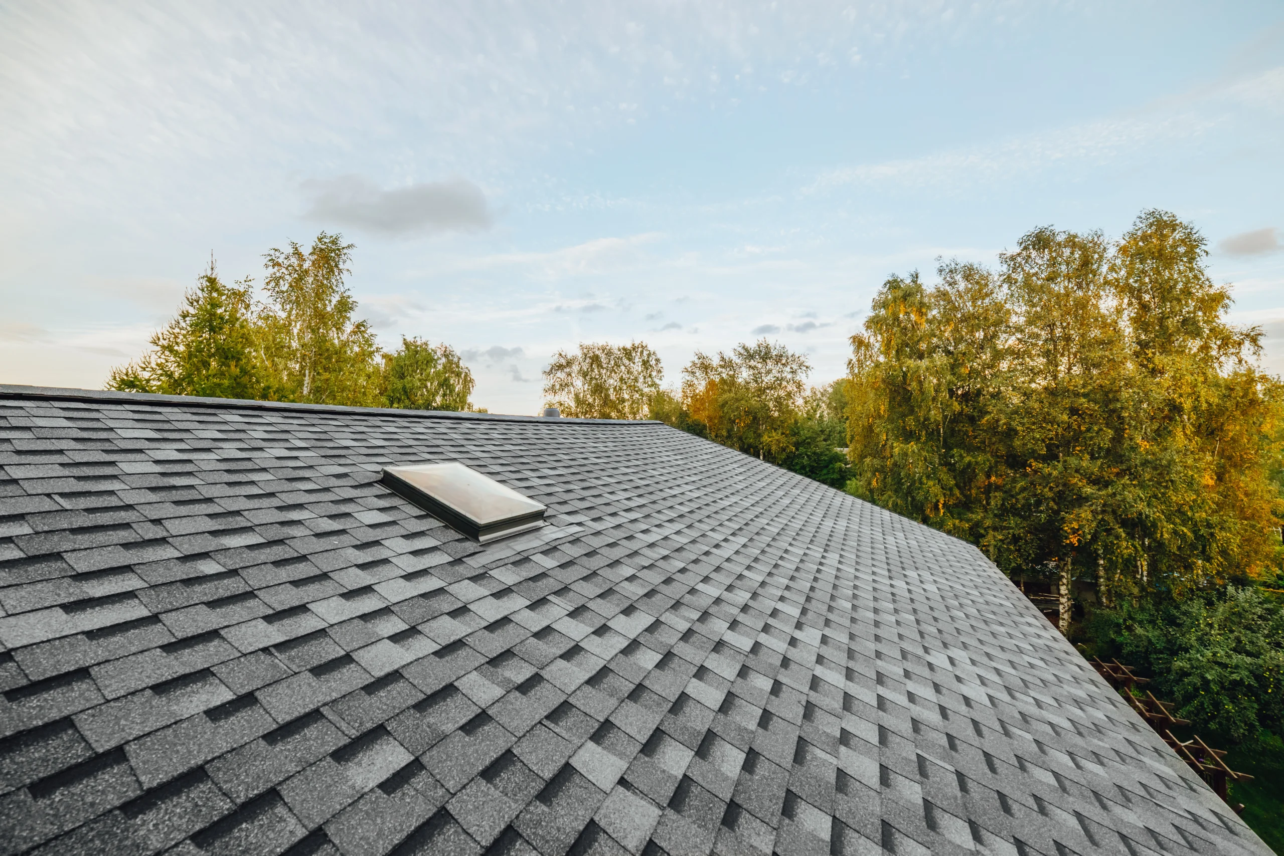 A close-up view of a gray shingled roofing job by Exterior Done Best, featuring a small skylight and surrounded by green and yellow trees under a partly cloudy Twin Cities sky.